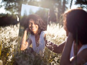 A woman sitting in a field of flowers holding a mirror & smiling at her reflection. If you struggle with body image concerns, reach out to begin therapy for women in NYC, NY. As a women's therapist, I am here to help!